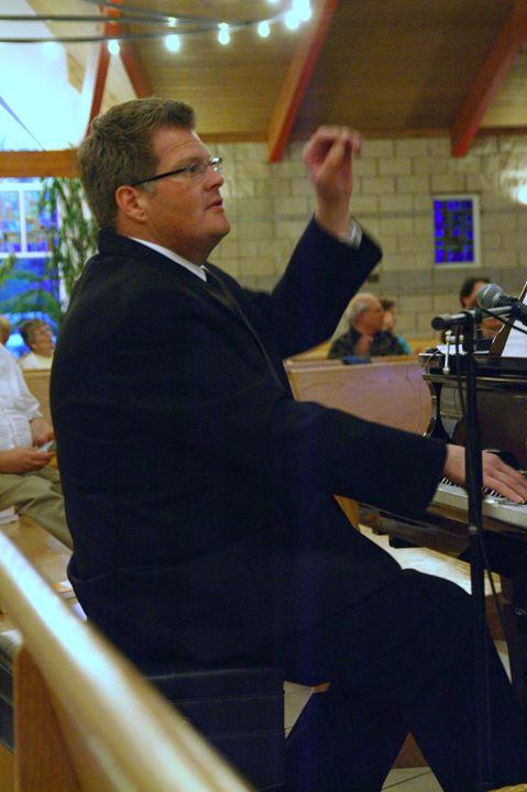 Dan Greig sitting at a piano, conducting a choir